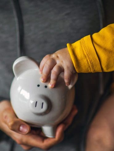 A baby holds on to the ear of a piggy bank while being held by its mother.
